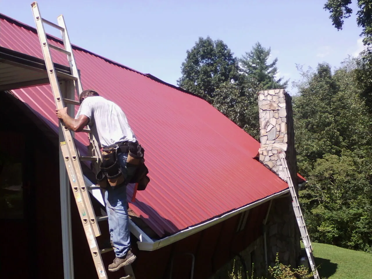 Roofer on a ladder beside a red metal roof and stone chimney — Soffit & Fascia Repair by Roofing Brothers in Valparaiso